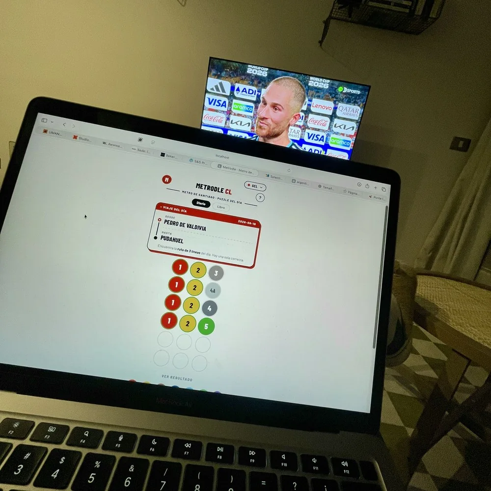 Metrodle CL abierto en un portátil de noche, con cuatro intentos ya jugados: cuatro filas de tres fichas de línea, y en la última la ficha verde de la línea 5 en su posición. Al fondo, un televisor con un partido