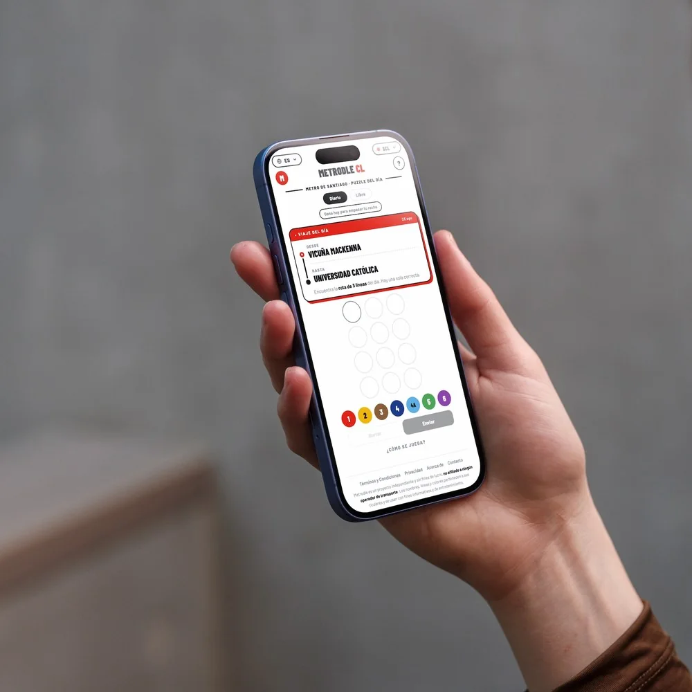 Una mano sostiene un teléfono con Metrodle CL abierto: el viaje del día va de 'Vicuña Mackenna' a 'Universidad Católica' y abajo espera la grilla de intentos